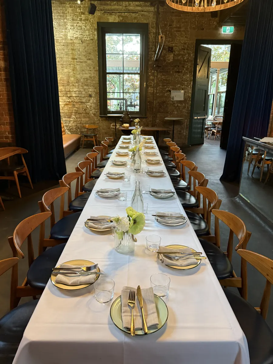 Long white-clothed table set for a dinner, with brick walls and a tall window